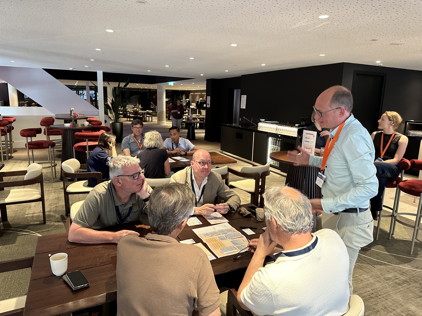 Five people seated around a table with the game laid out between them, while a facilitator stands and talks them through it.