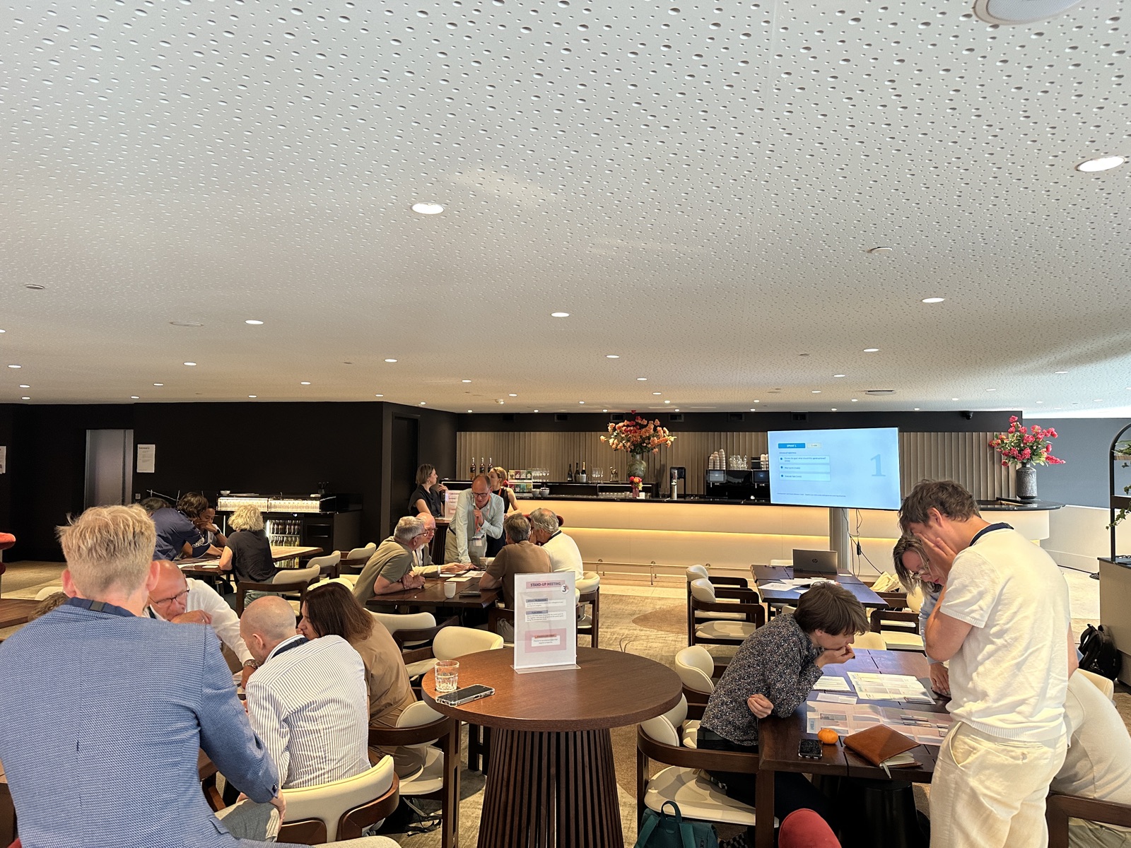 A hotel lounge at the Toezichtfestival, with several groups seated around tables working through the game and a presentation screen behind the bar.